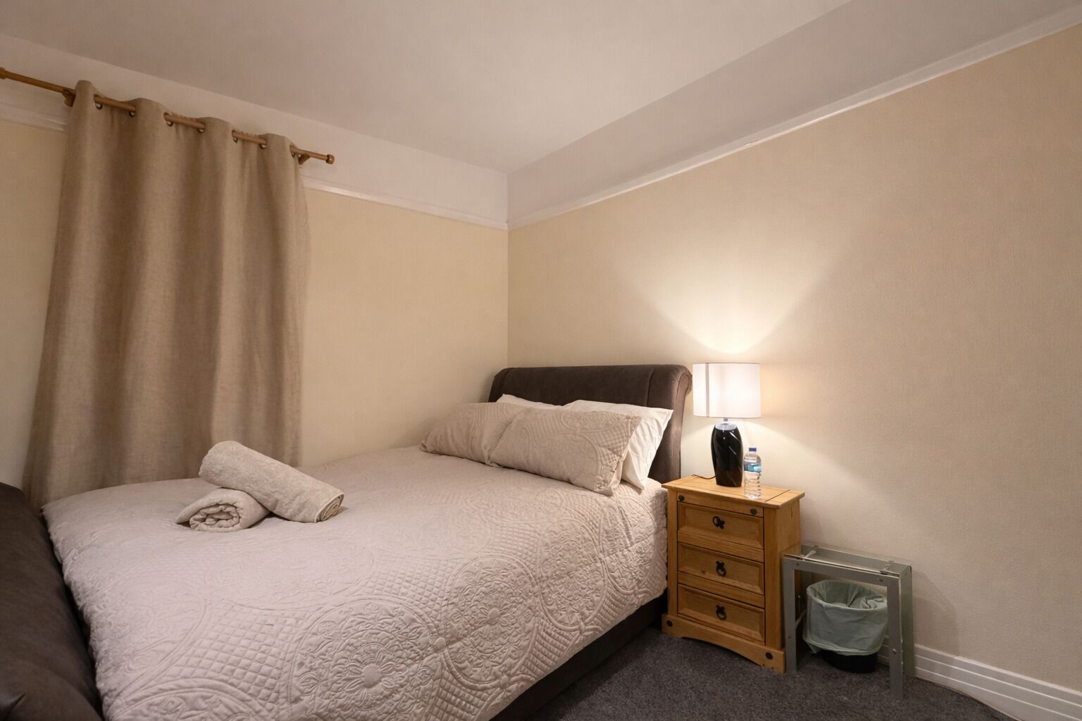 Una cama bien hecha con un edredón color crema, una lámpara de noche, una mesita de madera y un cubo de basura.