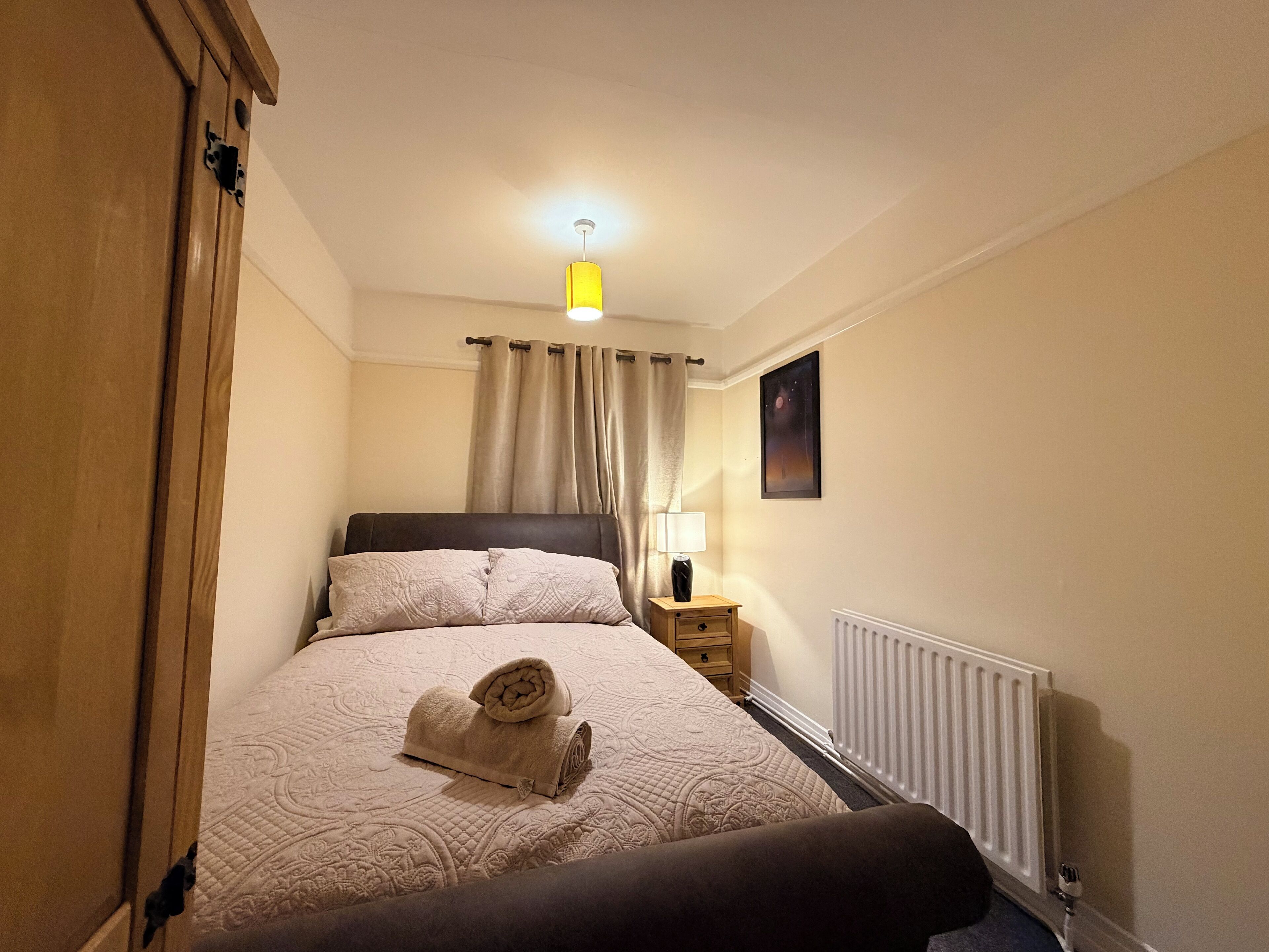 A bedroom with a bed, a nightstand, a lamp, a radiator, and a painting on the wall.