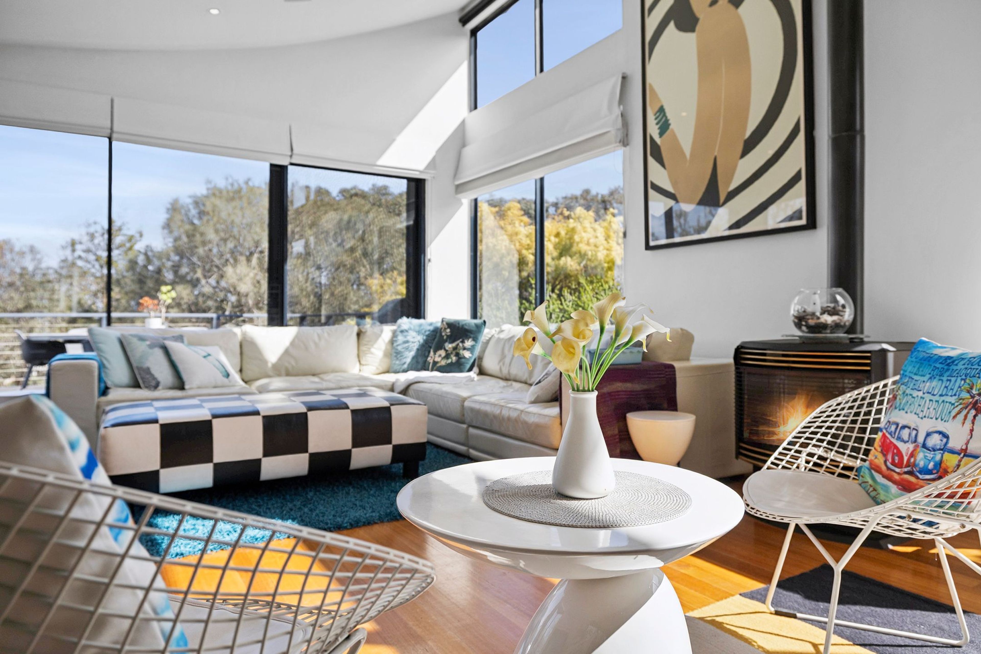 A modern living room with a checkered sofa, a round coffee table with a vase of flowers, and a large window offering a view of trees.