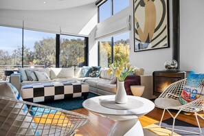 A modern living room with a checkered sofa, a round coffee table with a vase of flowers, and a large window offering a view of trees. - Blue Datcha (Aireys Inlet)