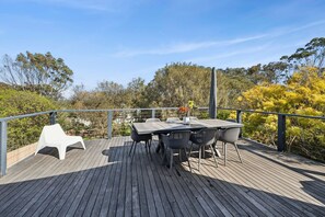 Eine Holzterrasse mit Tisch und Stühlen, umgeben von Bäumen und blauem Himmel.