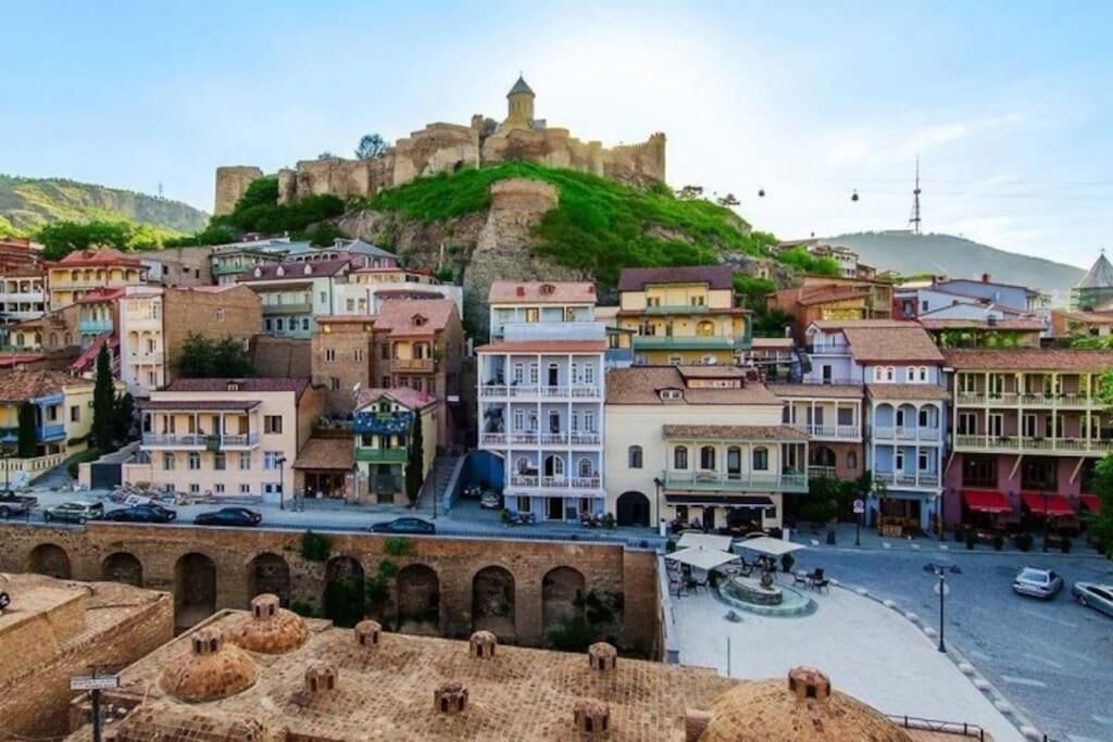 Un pueblo histórico con edificios coloridos, un castillo imponente en una colina y una plaza central con una fuente.