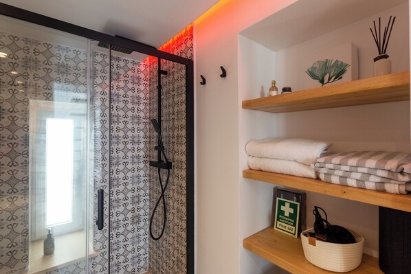 Een badkamer met een glazen douchedeur, een handdoekhouder met opgevouwen handdoeken, een verbanddoos en een plank met decoratieve objecten.