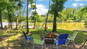 Een camping met een vuurkuil en stoelen, omringd door bomen en een meer.