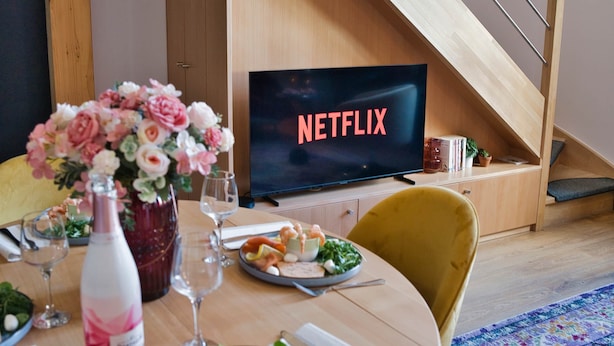 Ett modernt vardagsrum med en matplats, en tv-skärm som visar "NETFLIX", en trätrappa och ett matbord dekorerat med blommor och glas.