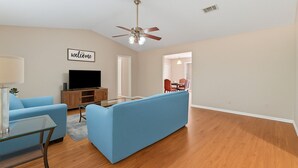 Un salon avec un canapé bleu, une table basse en verre, un téléviseur posé sur un meuble en bois et un ventilateur de plafond.