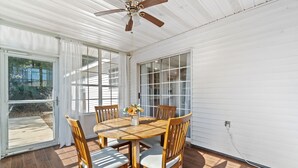 Une terrasse couverte avec une table et des chaises en bois, un ventilateur de plafond et de grandes fenêtres.