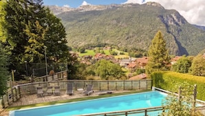 Una piscina con sillas de descanso, rodeada por una terraza de madera, con vistas a un pueblo con casas y montañas al fondo.