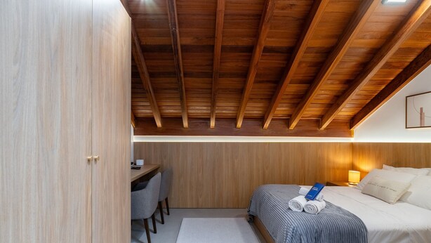 Un dormitorio con techo de madera, una cama con manta azul, un escritorio con silla y una mesita de noche con lámpara.