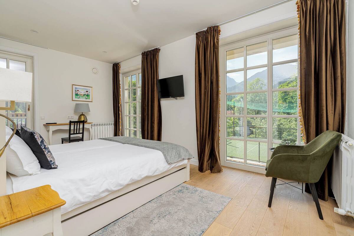 A bedroom with a bed, a desk, a chair, a TV, and a view of greenery through large windows.