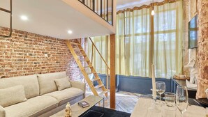 Ett rum i loftstil med synliga tegelväggar, en trätrappa, en soffa, ett matbord för fyra personer och ett stort fönster med tunna, genomskinliga gardiner.