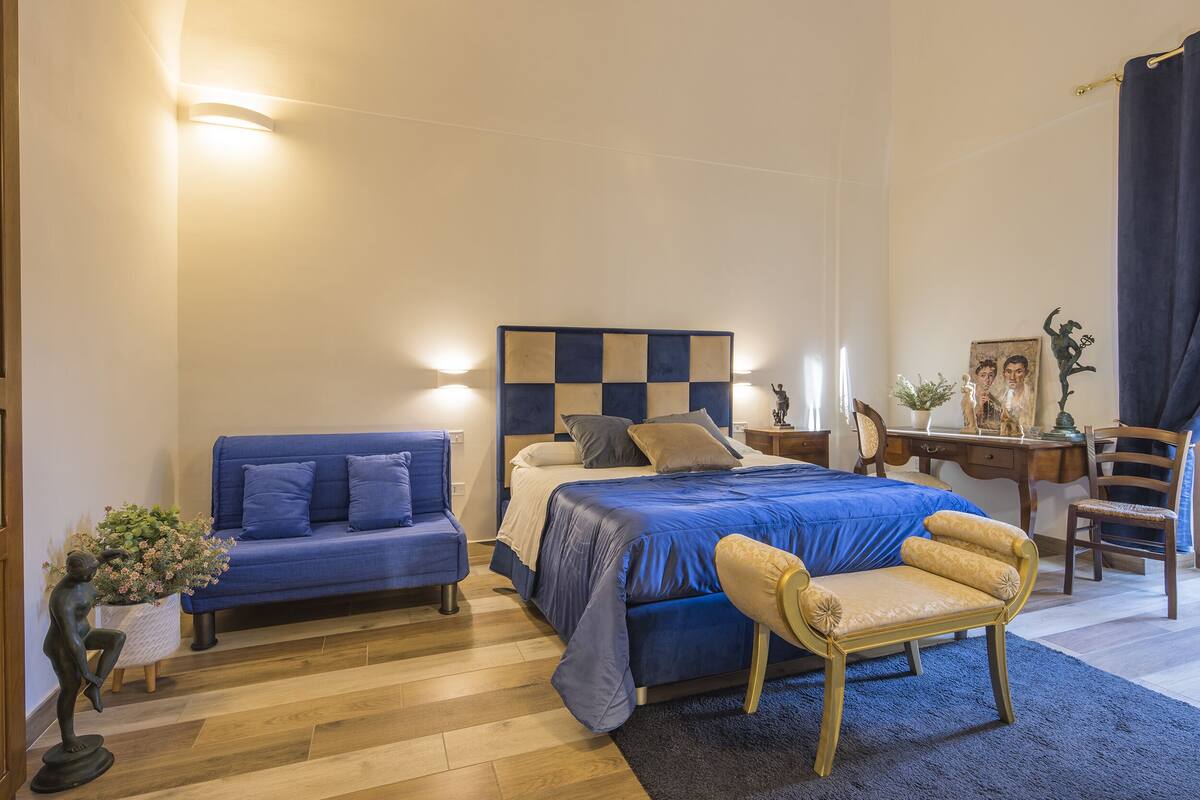A bedroom with a bed, a blue sofa, a wooden chair, and a desk with a lamp.