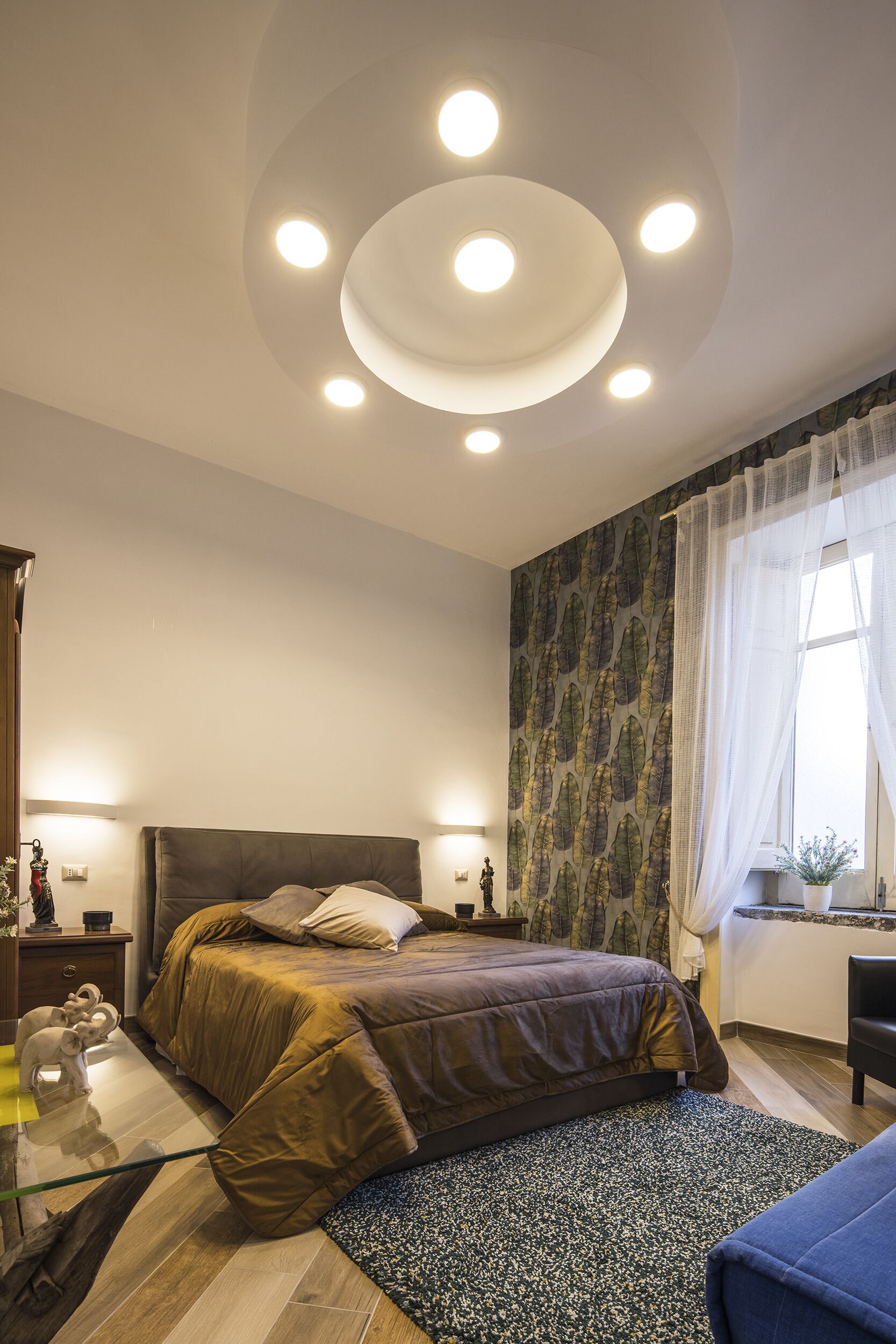 A bedroom with a bed, bedside tables, a nightstand, a window with curtains, and a ceiling with recessed lighting.