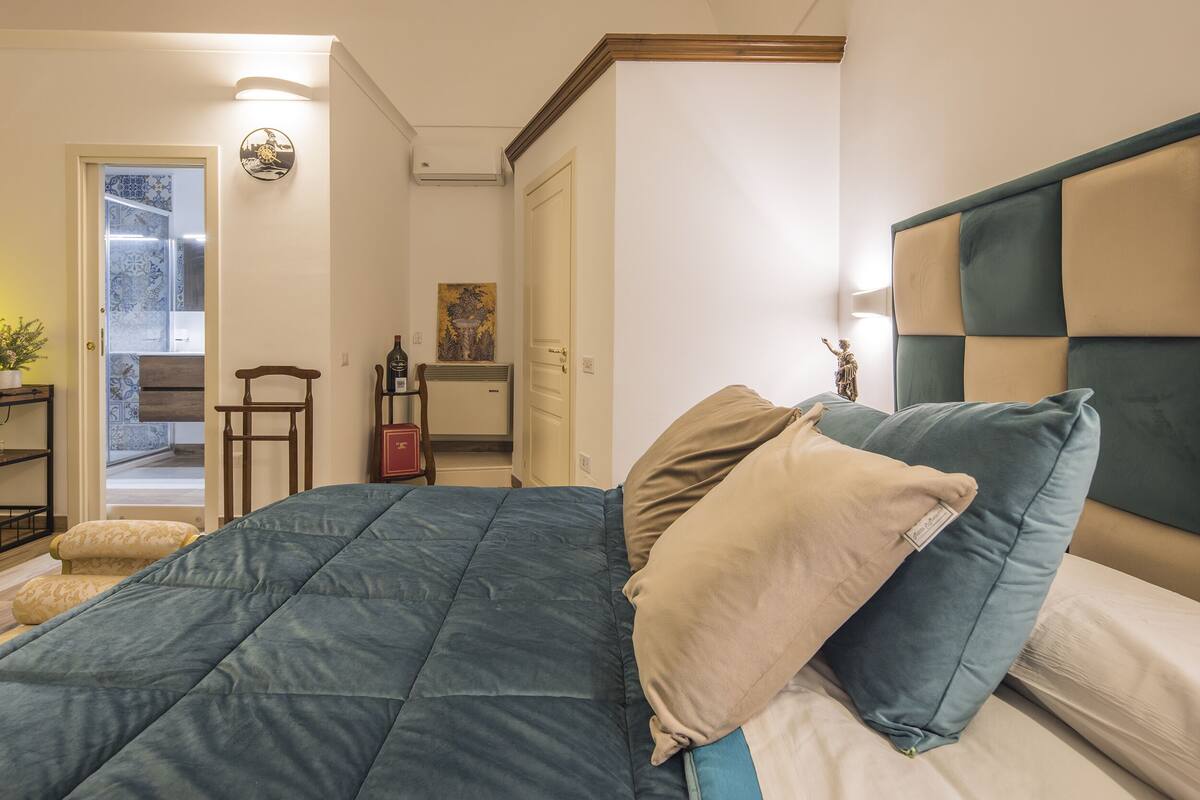 A bedroom with a bed, pillows, a nightstand, a lamp, and a door leading to another room.