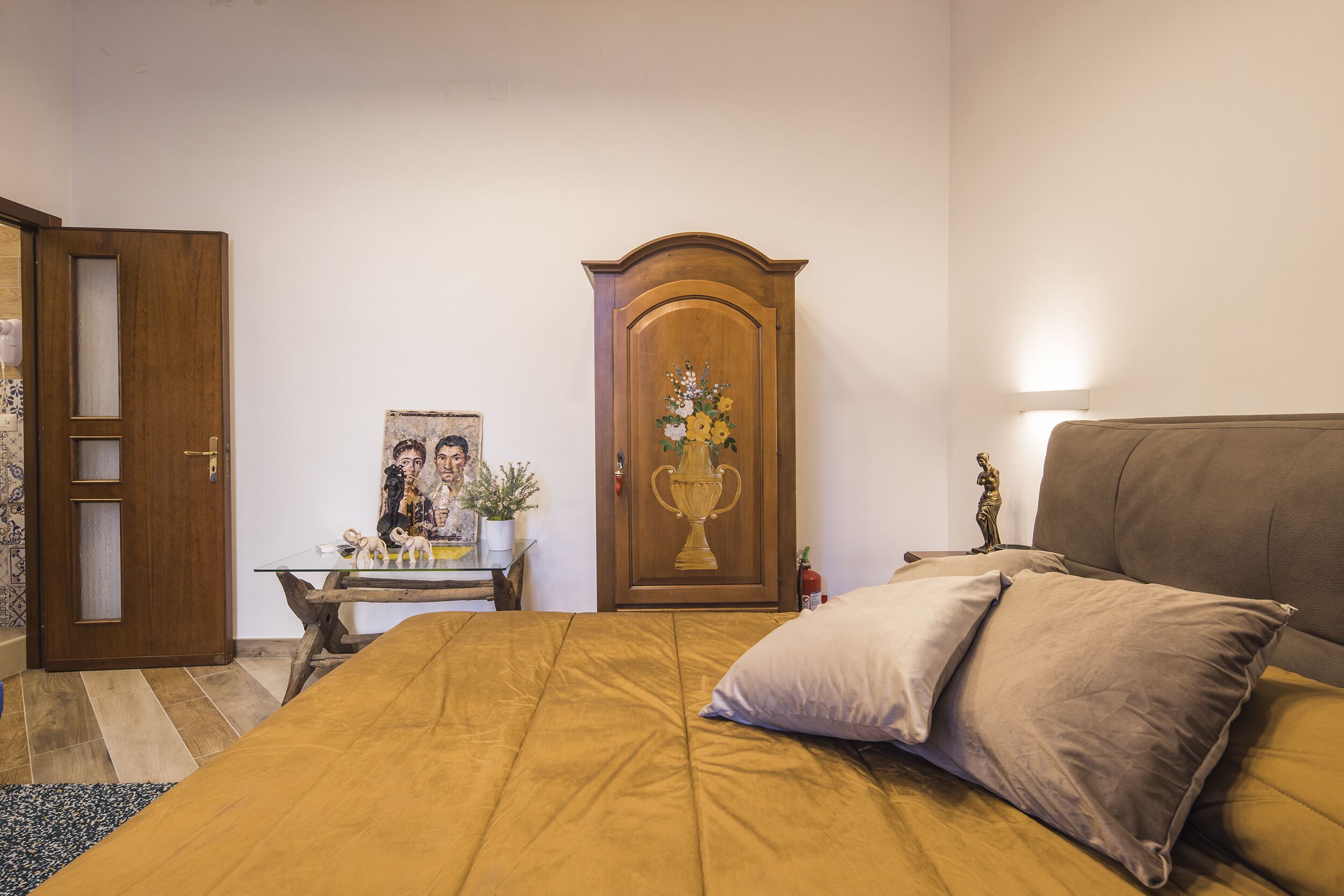 A bedroom with a bed, a wooden wardrobe, a small table with a vase, and a door.