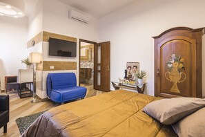 A bedroom with a bed, a blue chair, a TV mounted on the wall, a wooden door with a decorative handle, and a small table with a vase. - pompei suite via roma 16 (Pompei)