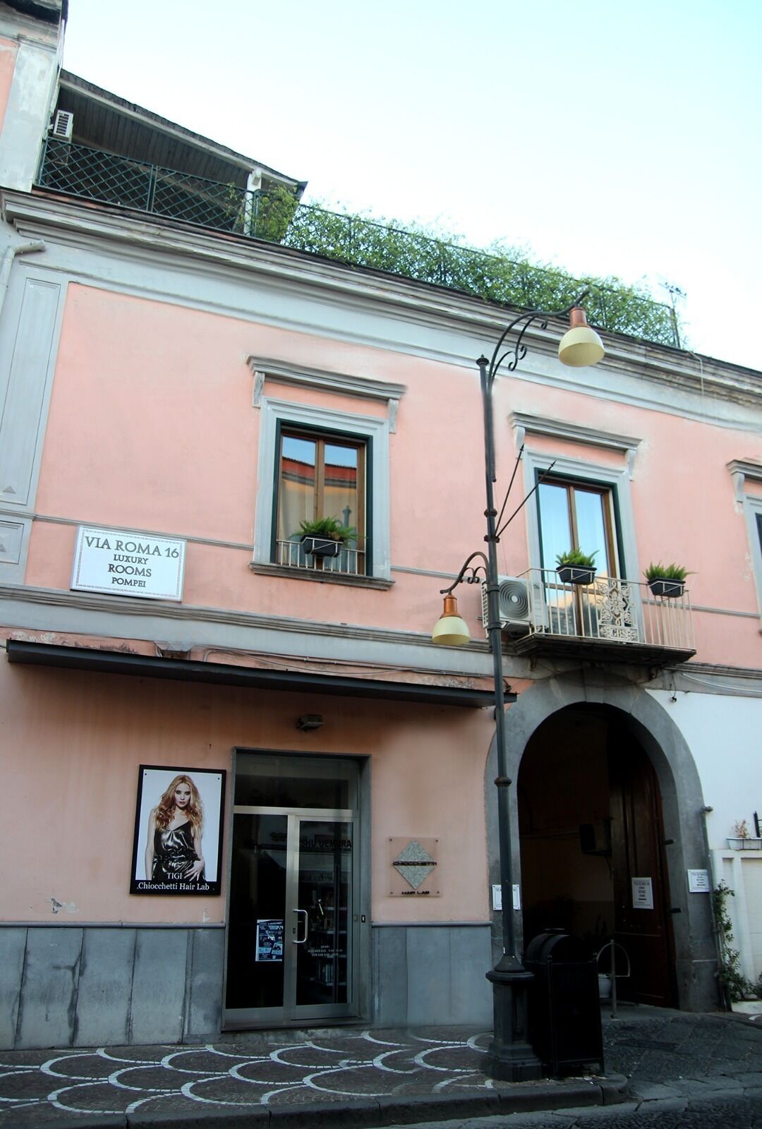 A pink building with a sign for "VIA ROMA 16 LUXURY ROOMS POMPEI" and a Chiocchetti Hair Lab.