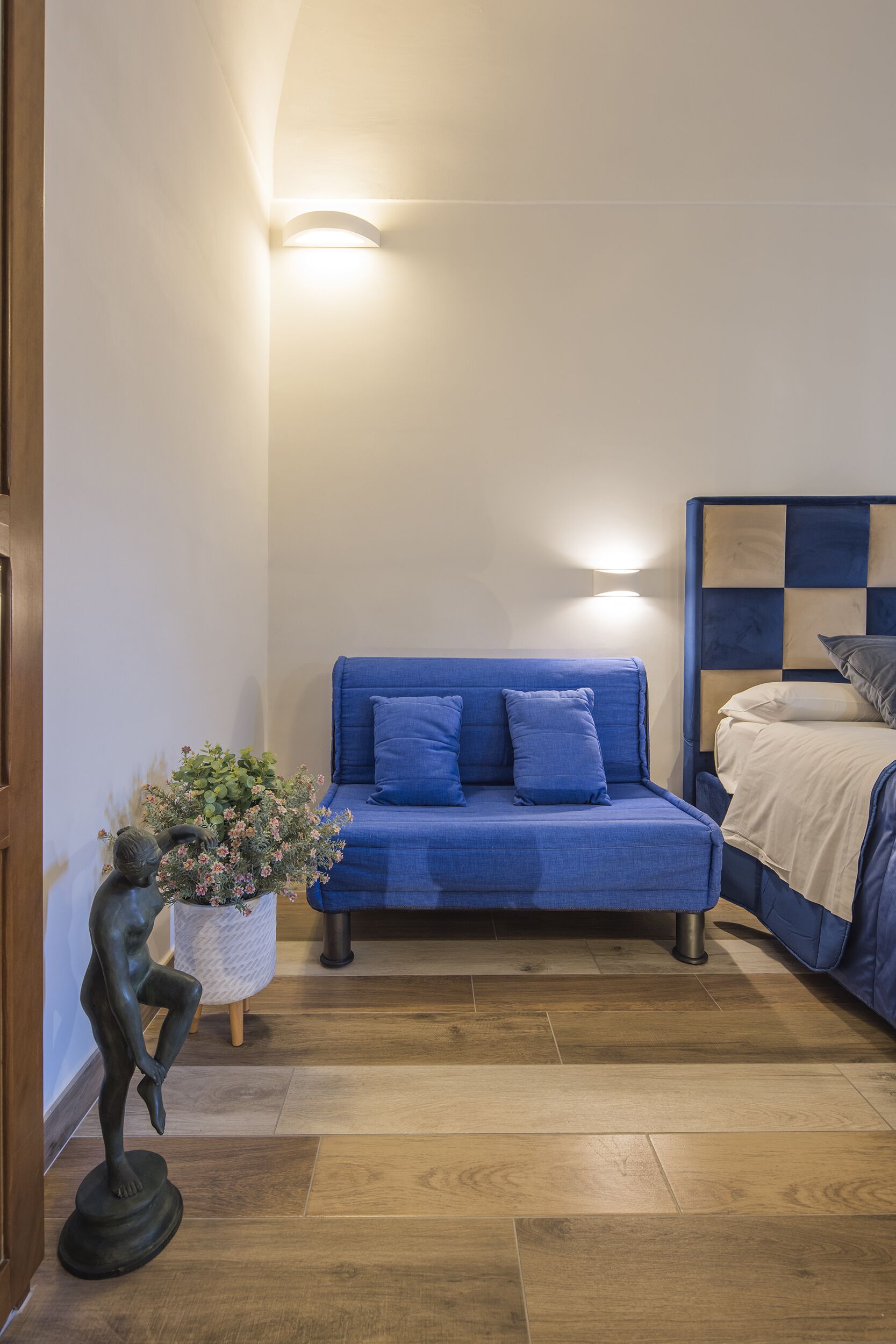 A bedroom with a blue sofa, a bed with a wooden headboard, a small plant, and a statue.