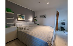 Een slaapkamer met een bed, nachtkastjes, een dressoir, een wandplank met decoratieve objecten, en een deur die naar een andere kamer leidt.