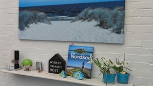 A beach scene painting on a wall, a book titled 'Die Nordsee,' and a shelf with decorative items. - Strandnaher Bungalow in Dornumersiel (Dornum)