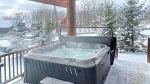 Un bain à remous sur une terrasse en bois, avec un sol enneigé, entouré d’arbres et de bâtiments.