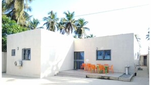 A small, single-story building with a flat roof and a concrete exterior. There are two windows with metal bars, a small air conditioning unit attached to the wall, and a set of outdoor orange chairs and tables.