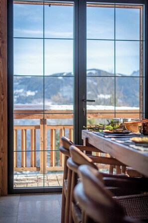 Une table à manger en bois avec des chaises, une vue sur les montagnes par une porte vitrée et une terrasse en bois à l’extérieur.