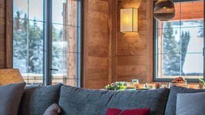 Ein gemütliches Blockhauskamin-Interieur mit Holzbalkendecke, großen Fenstern, einer Couch mit Kissen und einem Tisch mit Geschirr.