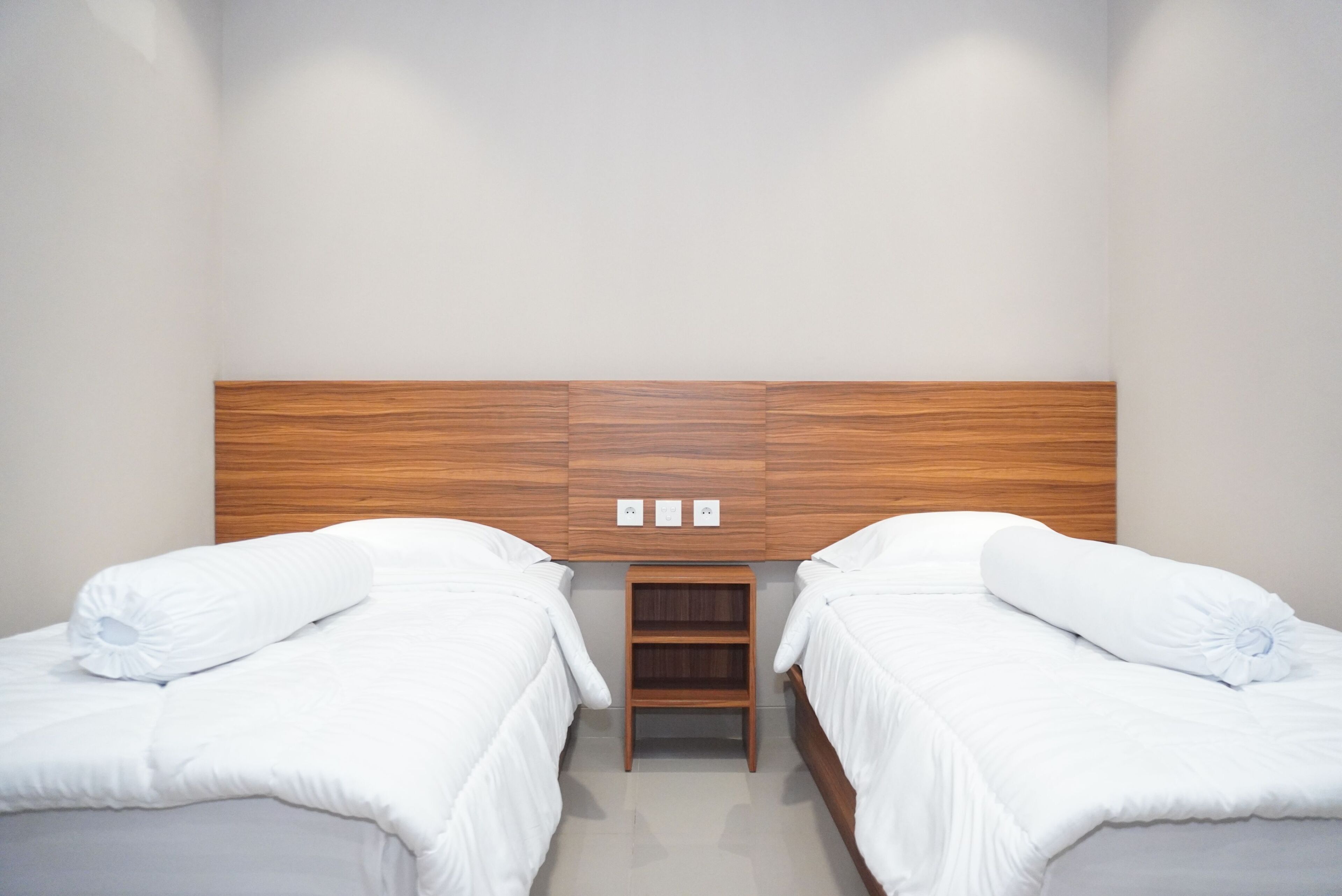 A hotel room with two single beds, a wooden headboard, a small wooden nightstand, and a wall-mounted power outlet.