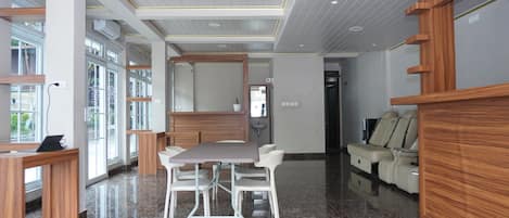 A modern hotel lobby with a marble floor, wooden reception desk, a seating area with white chairs, a sofa, and a large window offering a view.
