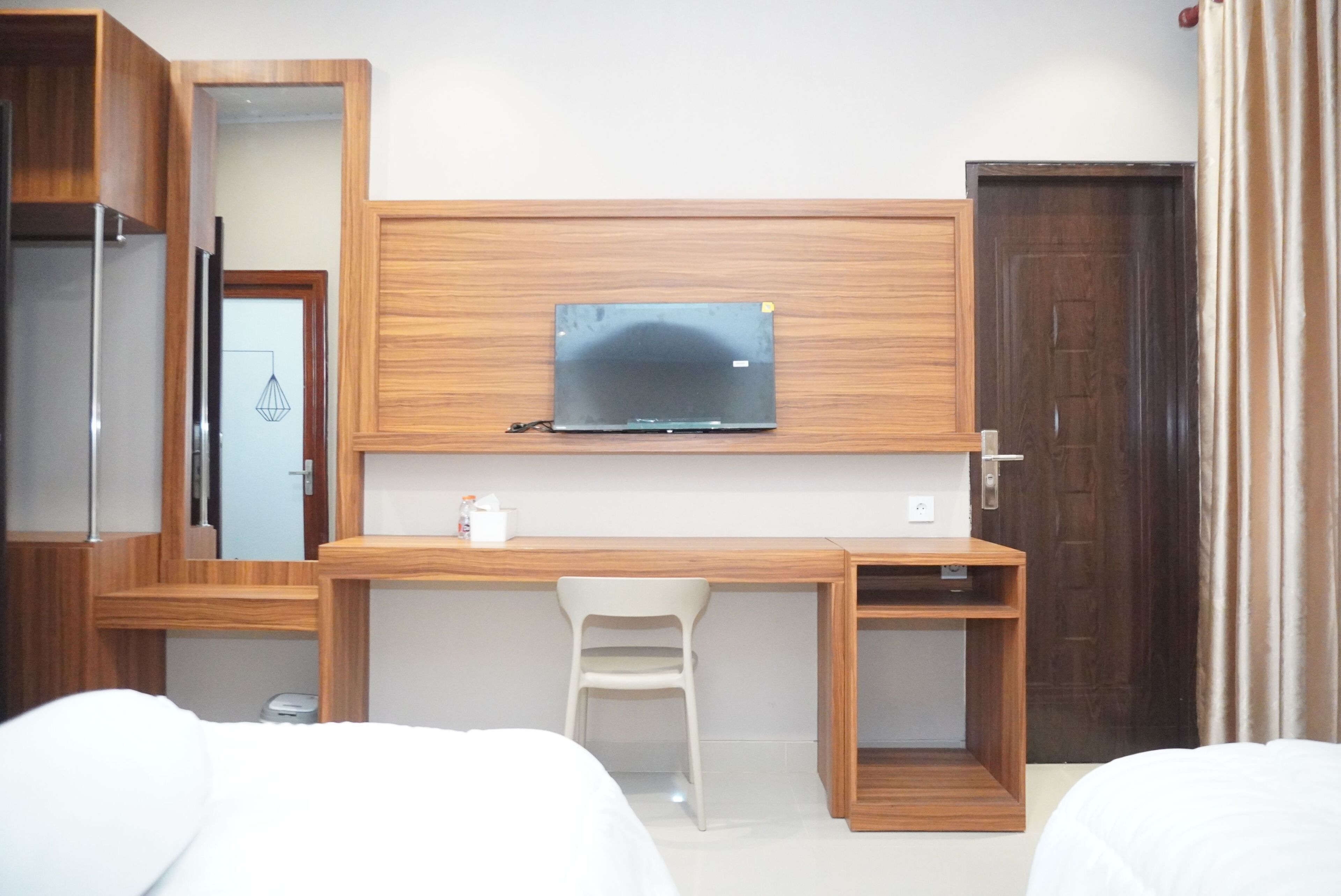 A hotel room with a wooden desk, a television mounted above it, a white chair, and a bed with white bedding.