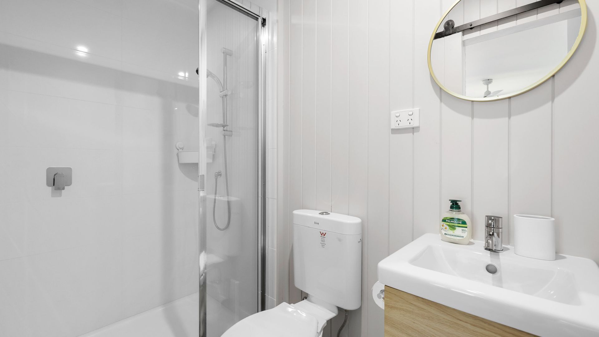 A modern bathroom with a glass-enclosed shower, a toilet, a sink with a mirror, and a bottle of Palmolive soap.