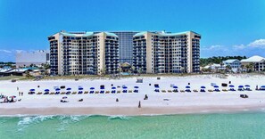 A beachfront hotel with multiple high-rise buildings, a sandy beach with sun loungers and umbrellas, and clear turquoise waters. - Beachfront Gulf Views | Beach Chairs Service + Pools (Panama City Beach)