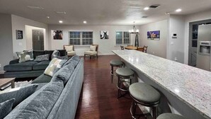 A modern living room with a grey sofa, a dining area with a bar, and a kitchen with a refrigerator.