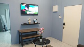 Ein Zimmer mit einem an der Wand befestigten Fernseher, einer Holzbank, einem kleinen Tisch mit Blumen in einer Vase und einer weißen Tür.