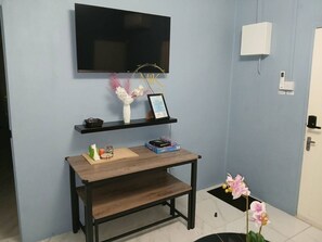 Una habitación con televisión de pantalla plana montada en la pared, un escritorio de madera con un marco de fotos, una pequeña estantería con adornos y una orquídea en maceta.