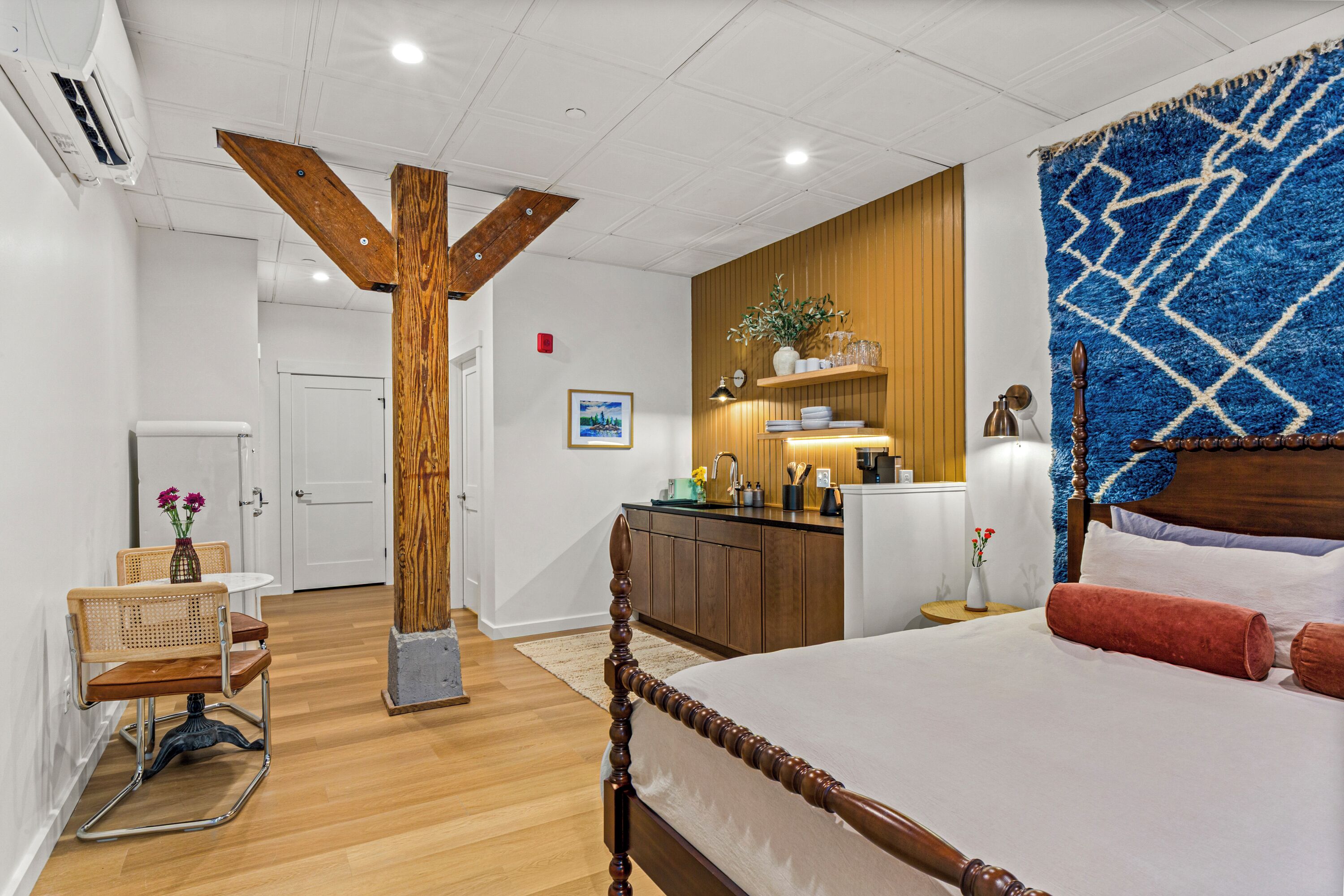 A hotel room with a wooden floor, a bed with a wooden headboard, a small table with a chair, a kitchenette, and a bathroom.