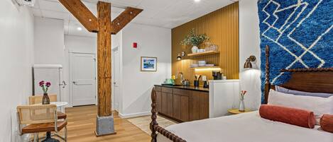 A hotel room with a wooden floor, a bed with a wooden headboard, a small table with a chair, a kitchenette, and a bathroom.