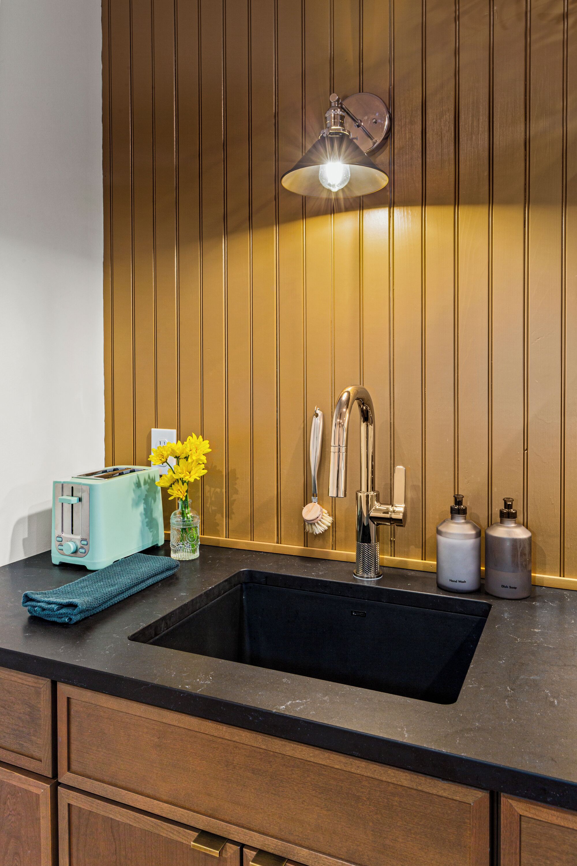 Eine Küche mit einem schwarzen Spülbecken, einem Toaster, einer Vase mit gelben Blumen, einem Wasserhahn und einer Wandleuchte.