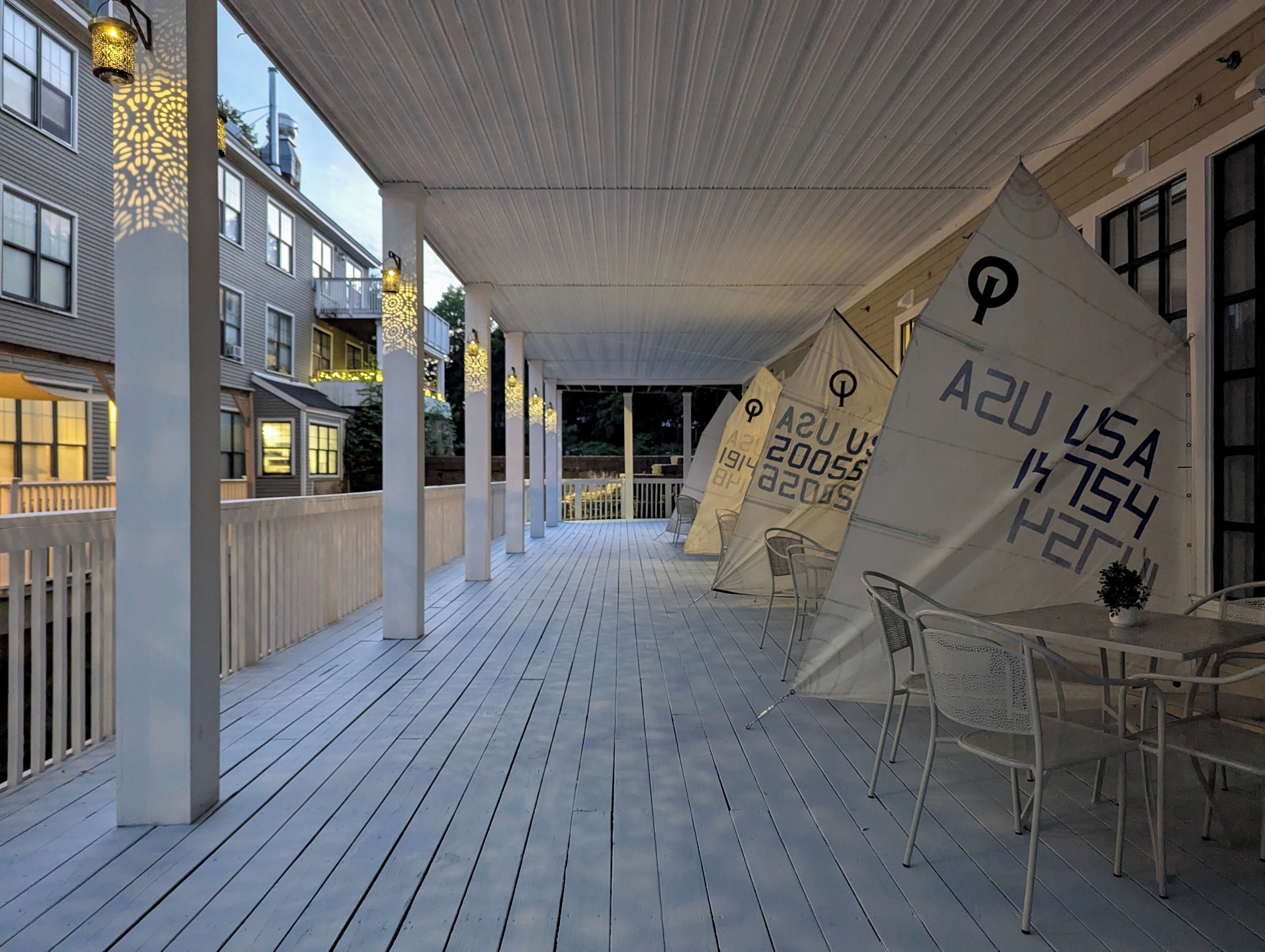 A covered outdoor area with a wooden deck, white columns, and a row of chairs.