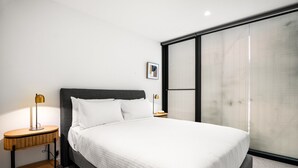 A modern hotel room with a large bed, a bedside table with a lamp, and a large window with blinds. - Stylish 3-bed Townhouse in Vibrant Fitzroy (Melbourne)