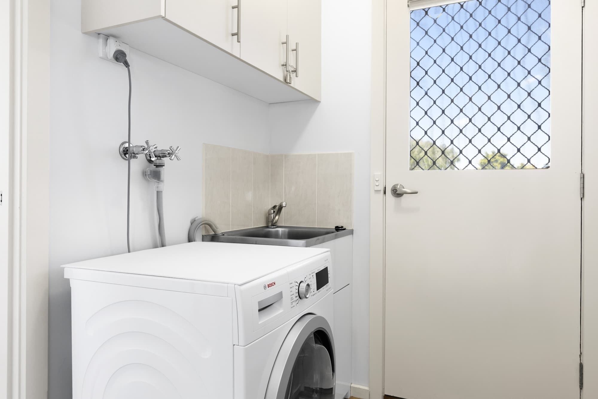 A compact laundry room with a Bosch washing machine, a sink, and a window with a mesh screen.