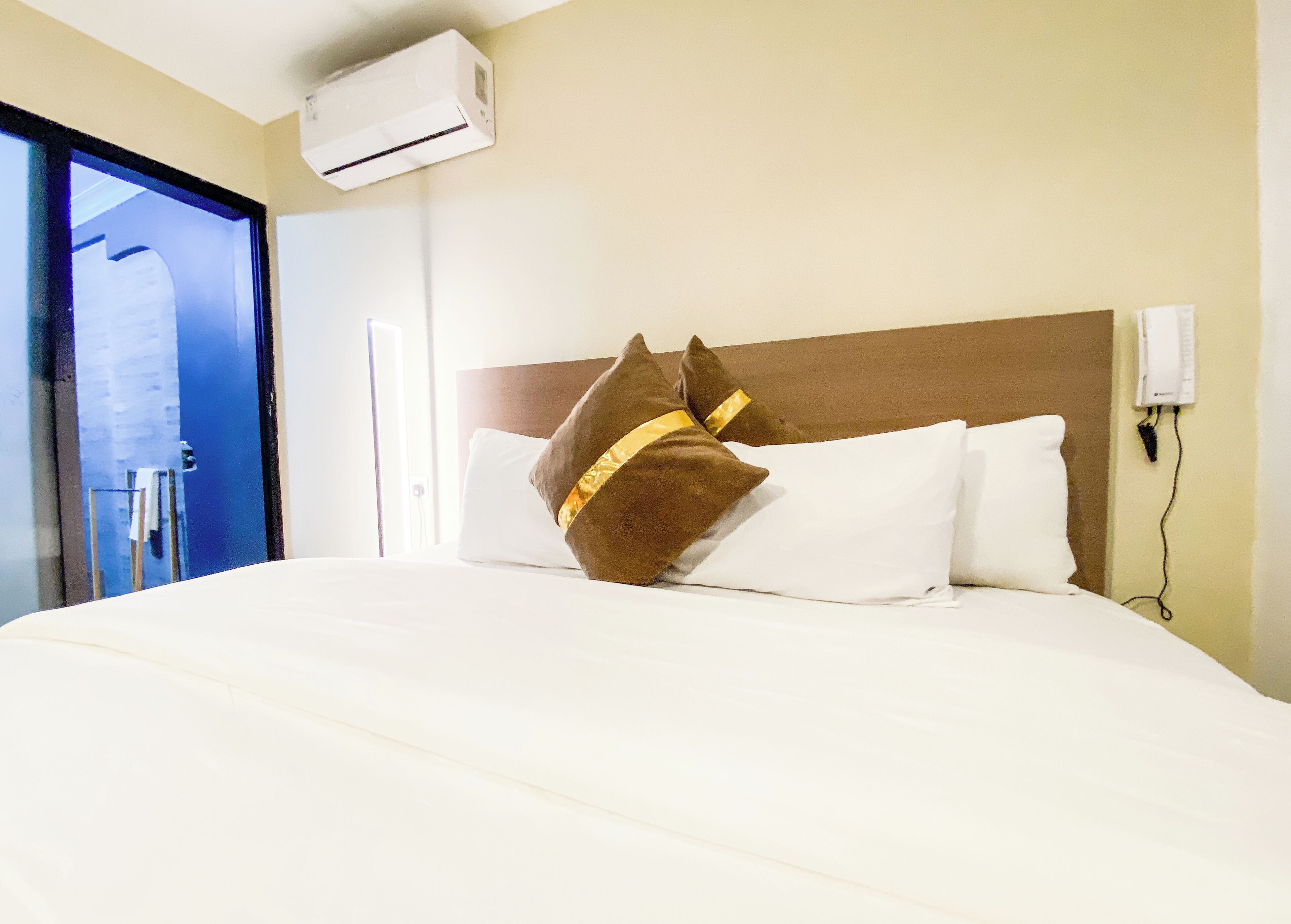 Habitación de hotel con cama, almohadas blancas, una almohada marrón con banda dorada, unidad de aire acondicionado, toma de corriente y puerta con panel de vidrio.