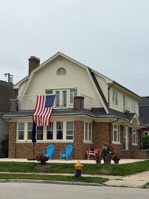 A two-story house with a prominent American flag on the front porch, a brick facade, and a small front yard with potted plants and outdoor furniture. - Lake house North Beach lake breezes, morning sunrises and evening sunsets (Racine)