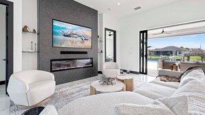 En moderne stue med et fladskærms-tv, en pejs og udsigt til et poolområde og et hus.