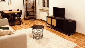 Un salon moderne avec un plancher en bois, un canapé, une table à manger avec des chaises, un meuble pour la télévision, une bibliothèque noire et des œuvres d’art encadrées accrochées au mur.
