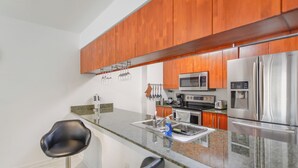A modern kitchen with wooden cabinets, stainless steel appliances, granite countertops, and two black bar stools. - Brickell Bay 2308 (Miami)