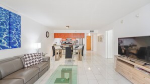 A modern living room with a sofa, coffee table, and TV. A kitchen with orange cabinets is visible in the background. - Brickell Bay 2308 (Miami)