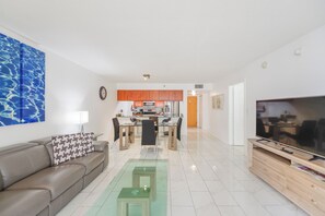 A modern living room with a sofa, coffee table, and TV. A kitchen with orange cabinets is visible in the background. - Brickell Bay 2308 (Miami)
