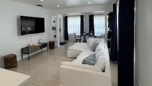 A modern living room with a white sofa set, a flat-screen TV mounted on the wall, and a dining area in the background. - 2BR/1B - Sunny Hollywood Hideaway (Hollywood)